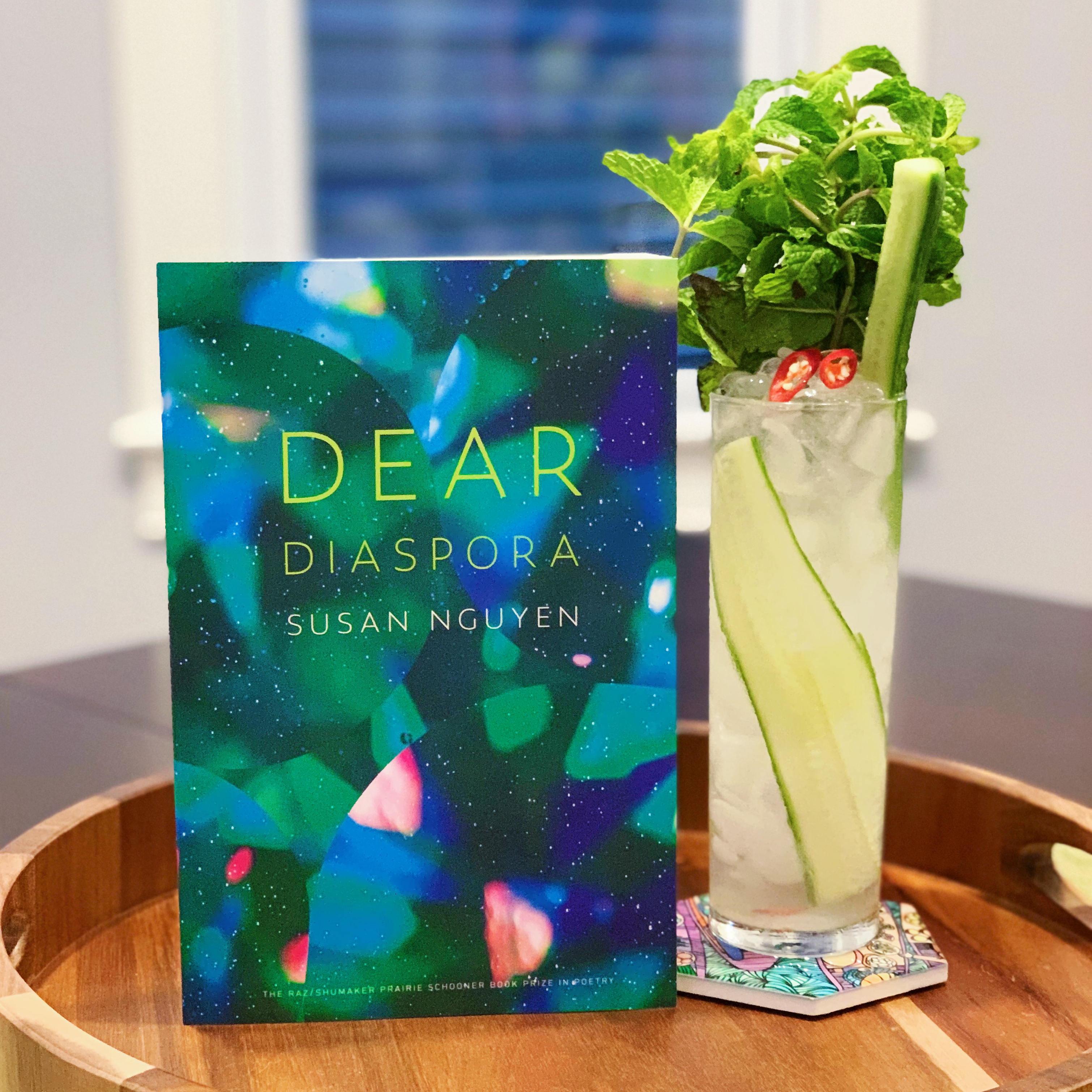 book titled Dear Diaspora, propped next to a cocktail in a tall glass garnished with mint, cucumber, and Thai chili pepper